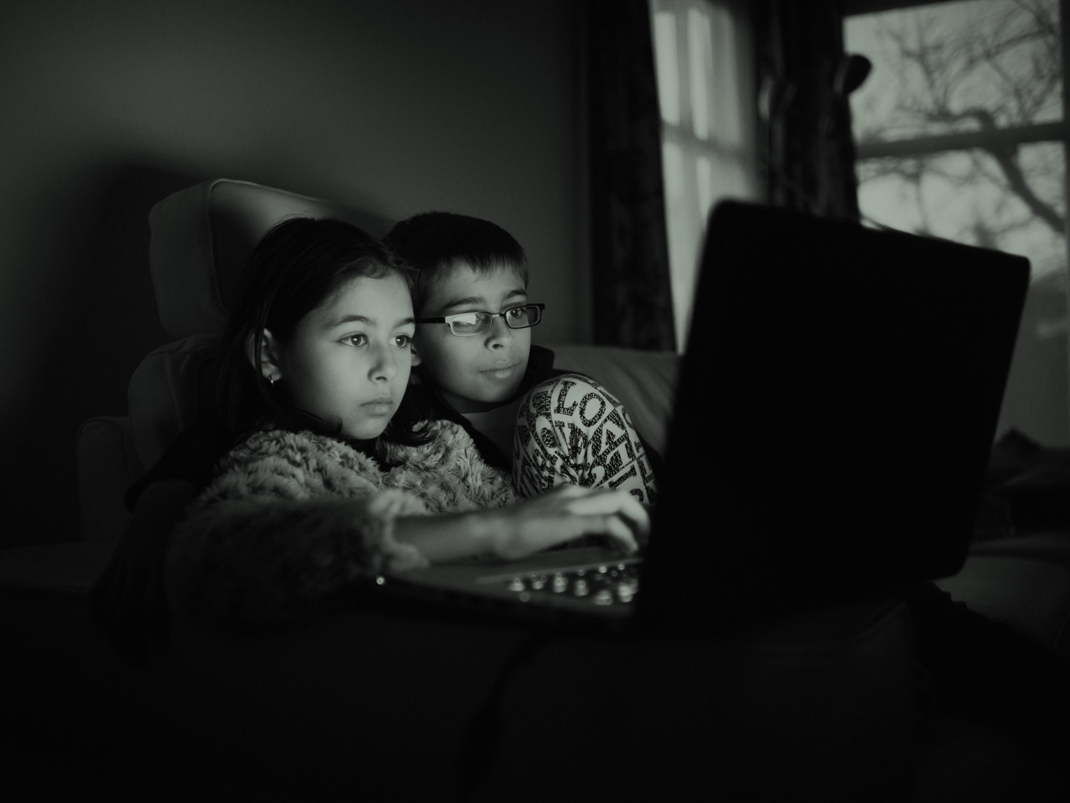 Duas crianças atrás de um laptop no escuro, iluminado por uma tela de computador